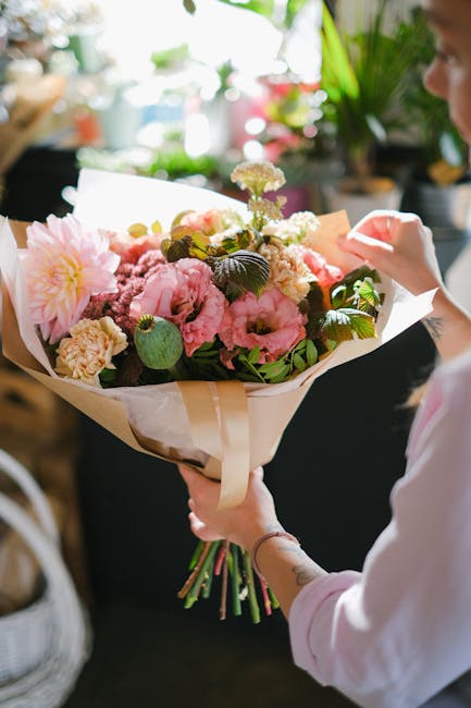 A person holding a large floral bouquet featuring soft pink peonies, white carnations, and small purple blossoms, wrapped in cream-colored paper with a satin ribbon. The bouquet is arranged with lush 