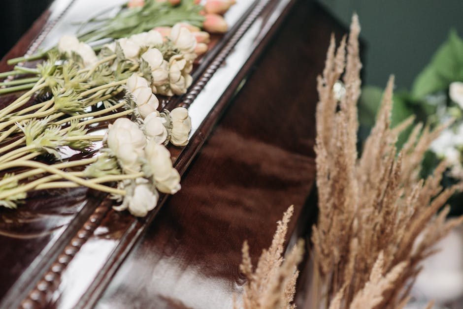 A floral tribute featuring a delicate arrangement of white roses and pale pink carnations laid on a polished wooden surface, with visible stems and petals that appear fresh and soft in texture. The fl