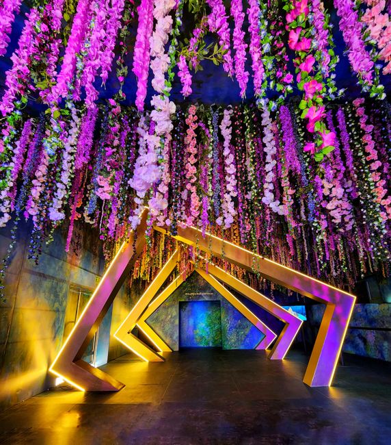 A vibrant floral installation featuring cascading purple, pink, and lavender flowers hanging from the ceiling, creating a lush, colorful canopy. Beneath the floral display are modern geometric archway