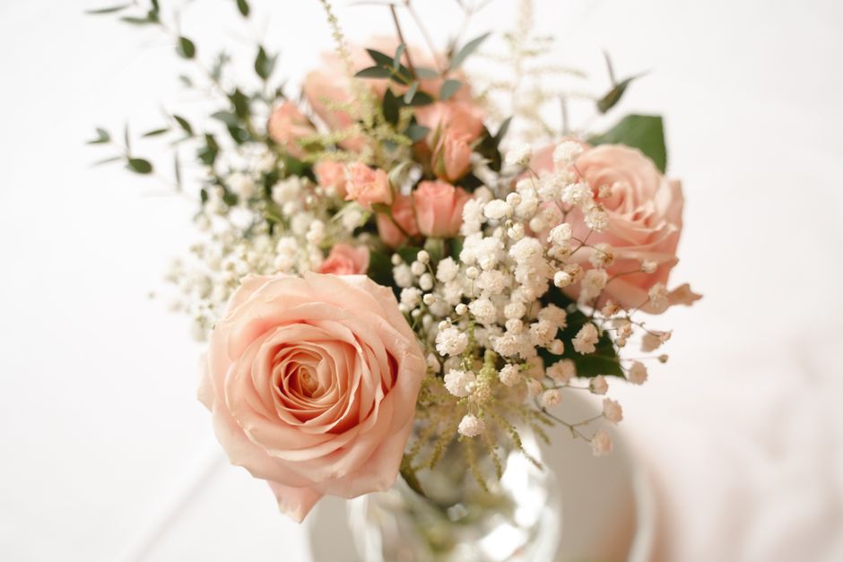 A close-up of a floral arrangement featuring a large, fully bloomed pale pink rose with softly layered petals, surrounded by smaller pink roses still in bud form. The bouquet includes delicate white b