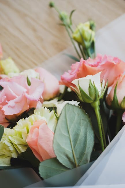 Close-up of a floral gift comprising soft pink and cream lisianthus blossoms with delicate, ruffled petals, complemented by fresh green leaves and stems. The bouquet is partially wrapped in white tiss