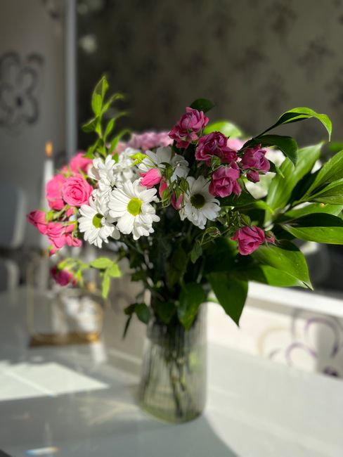 A clear glass vase containing a fresh flower bouquet positioned on a white table. The arrangement includes white daisies with yellow centers, pink roses, and small pink flowers, complemented by lush g