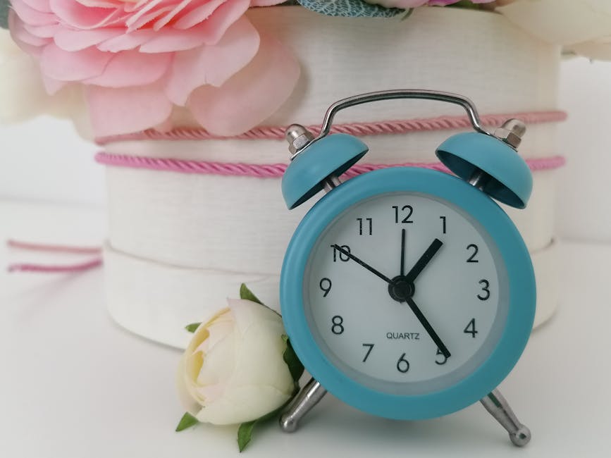 A light blue vintage-style alarm clock with twin bells and a white face displaying the time as 12:03, placed on a white surface. Next to the clock, there is a delicate white rose with soft petals and 