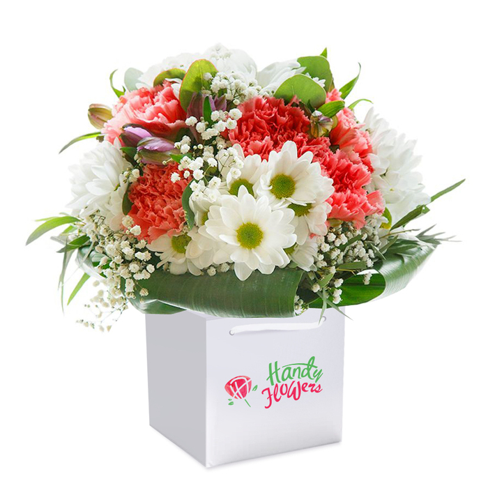 Expertly arranged by trusted local florists, this exuberant bouquet sits in a crisp, square white gift bag, showcased against a clean, radiant background that lets every bloom shine. The core is formed by vibrant coral and peach carnations, their countless ruffled petals layered to create a plush, inviting texture. Cheerful white daisies, set apart by their citrus-green centres, peek from between the larger flowers, forming a playful counterpoint that reflects the optimism of Stevenage's local neighbourhoods. Accents of baby's breath float gracefully through the bouquet, their tiny blossoms imparting both lightness and a gentle, modern touch. Subdued but intriguing, purple-tinged, unopened buds add a note of growing anticipation, mirroring the excitement of community celebrations at places like Stevenage Arts & Leisure Centre. Broad leaves of glossy, deep green form a natural base, wrapping the bouquet and grounding the colourful exuberance above. The white ribbon handles lend style and practicality, made for easy gifting-be it a same-day delivery to a friend near St Nicholas or a heartfelt gesture for a loved one in Chells. Clear, tasteful branding on the bag nods to Flowers Stevenage's reliability, while the entirety of the bouquet radiates freshness, thoughtfulness, and an easy elegance suited to any special event in Stevenage.
