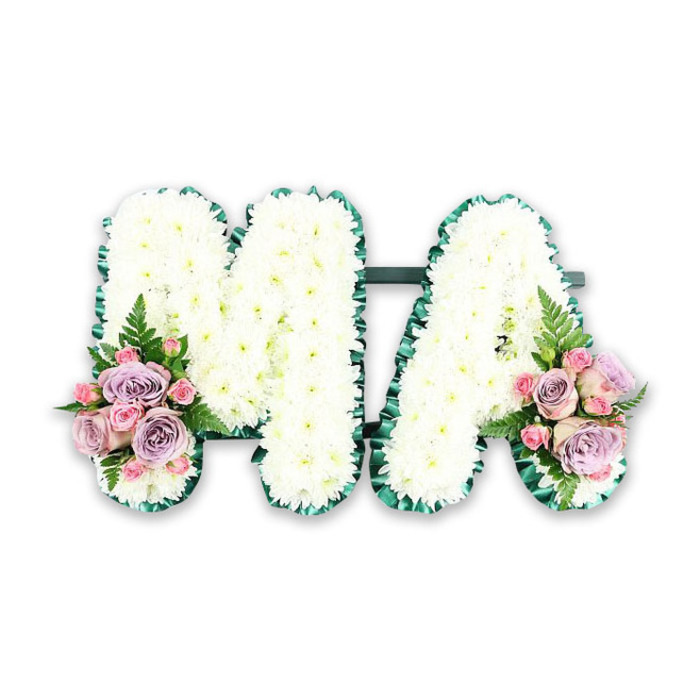 A personalised funeral flower tribute spelling the letters MA is displayed against a crisp white background, making the design appear calm, uncluttered, and dignified. The two capital letters are solidly constructed, then completely covered with compact white chrysanthemums that sit shoulder to shoulder, leaving no space between them. Each bloom adds its own ring of fine, slightly curved petals, together creating a thick, quilted texture that looks pillowy and comforting. The white is gentle rather than stark, softened by the natural form of the flowers. Tracing the outside edge of both letters is a deep green ribbon, likely satin, carefully pleated to form a continuous, ruffled frame. This border adds definition and a touch of formality, guiding the eye cleanly around the shape of the M and the open centre of the A. At the lower left of the M and the lower right of the A, matching accent sprays of flowers bring colour and emotional warmth. These clusters are composed of muted lilac roses and pale pink roses, interwoven with smaller pink spray roses that tuck into the spaces between larger blooms. The roses are at mixed stages of opening, revealing layered, spiralling petals that look velvety and delicate. Fern fronds add a feathery green lift, creating a contrast in shape and texture against the rounded roses and cushioned chrysanthemums. The tribute feels intimate and heartfelt, exactly the kind of bespoke piece a family in Stevenage might choose for a funeral at a local church or a service near the Old Town, where personal touches matter. The MA letters suggest a loving message for a mother, mama, or someone with those initials, with the calm whites and gentle pastels expressing remembrance, love, and a quietly spoken goodbye.