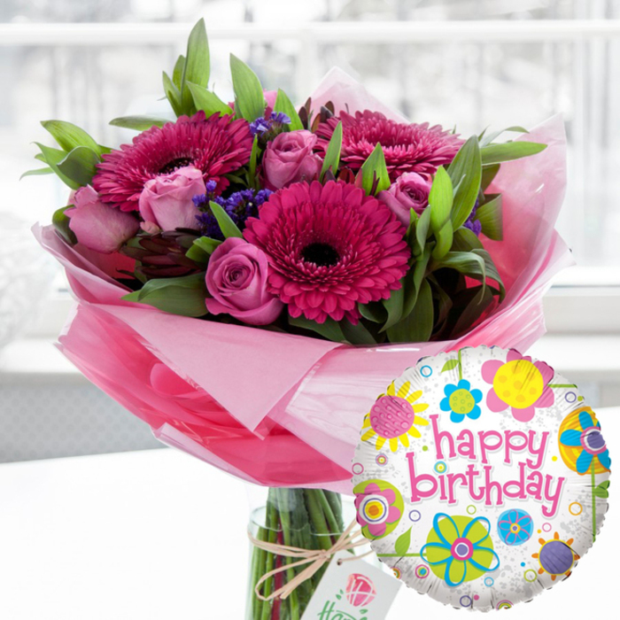An exuberant birthday gift set is shown as if just placed on a table near a sunny window in a Stevenage home, combining an elegant flower arrangement with a colourful foil balloon. The central focus is a lavish bouquet in a clear glass cylinder vase, the stems neatly aligned and visible through the water for a fresh, just-cut look. Around the neck of the vase, rustic twine is tied in a simple bow, holding a small off-white tag printed with a refined rose logo, giving the impression of a carefully curated, premium bouquet rather than something mass-produced. The flowers themselves form a rich tapestry of pinks and purples. Large gerbera daisies in intense fuchsia and magenta shades stand proudly, their almost black centres drawing the eye inward. Nestled among them are clusters of blush and pastel pink roses, some still in tight buds, others partially unfurled to reveal layers of soft, velvety petals. Here and there, delicate sprays of deep purple filler flowers, likely statice, peek through, adding depth and a cooler note to balance the warm tones. Lush green foliage, with both broad leaves and finer sprigs, surrounds the blooms, creating a rounded, abundant shape reminiscent of the greenery you might see in landscaped gardens around Great Ashby. The base of the bouquet is wrapped in a soft, translucent pink cellophane that flares lightly, adding a romantic, feminine touch and a gentle sheen. In the lower right foreground, overlapping the bouquet, a round Mylar balloon adds a burst of birthday fun. It has a white background with subtle grey speckles and is ringed with playful floral and circular patterns in sunshine yellow, lime green, bright blue, pink, and purple. Across the middle, the phrase happy birthday is written in bold pink script, echoing the gerberas' hue. The softly blurred indoor background and bright, natural light frame the entire scene as a warm, joyful celebration moment, ideal for marking special birthdays anywhere across Stevenage.