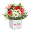 Radiating joy and tenderness, this luminous bouquet overflows from a pristine square white gift bag, perfectly poised for a special delivery anywhere in Stevenage. Plush coral and peach carnations, each petal edged in gentle ruffles, provide the foundation of warmth and fullness. Looming cheerfully throughout, pure white daisies with shining green-yellow centres add a playful brightness, their simplicity echoing the easy beauty of flower beds in Shephalbury Park. Wisps of white baby's breath weave delicately through the arrangement, gifting it an airy, almost dreamlike quality, while on the side, small, unopened purple-streaked buds bring an unexpected charm-hints of beauty yet to unfold. The bouquet is encircled by abundant glossy green leaves, their rich sheen adding structure and a flourishing freshness. Sturdy white handles arc above, ensuring this floral gift is as practical as it is elegant-ready to congratulate, celebrate, or comfort with a personal touch. Every flower is set with artistry and care, capturing the creative energy that defines Flowers Stevenage and the vibrant community it serves. This composition is more than just a gift; it's a radiant snapshot of shared happiness, perfect for uplifting any occasion from Stevenage Old Town to Chalkdene.
