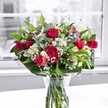 A lush, romantic bouquet of deep red roses and matching red carnations glows at the centre of the image, arranged with care in a classic clear glass vase set on a bright white table. Each rose, some fully open with velvety crimson petals and others still in tight buds, leans gently into the light, giving the arrangement a feeling of unfolding love and celebration. The carnations echo the same rich tone, their ruffled, full heads adding texture and old-fashioned charm, like something you might spot on a special anniversary table in a cosy Stevenage restaurant near the Old Town High Street. Threaded between the bold red blooms are crisp white alstroemeria, or Peruvian lilies, some still closed, others just starting to open so that their trumpet-shaped petals and soft green-tipped buds peek out shyly from the greenery. Tiny white gypsophila, or baby's breath, is scattered like a fine mist throughout, softening the contrasts and creating a cloud-like halo that gently brightens the composition. Around the base, thick, glossy green leaves form a strong collar, while more slender, lighter green foliage loops and weaves upward, adding movement and a feeling of freshness you might associate with a peaceful walk in Fairlands Valley Park. The clear vase reveals the neatly trimmed stems resting in clean water, a quiet reminder of the flowers' just-cut vitality, as though they've only just arrived from a local grower. Behind the bouquet, a large window is softly blurred, the view outside muted to grey and pale tones so that all attention stays on the vivid reds, whites, and greens. The overall mood is one of heartfelt appreciation and joy, perfect for a birthday, romantic surprise, or to brighten a home in Stevenage on any ordinary day that deserves to feel special.