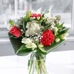 A contemporary mixed bouquet in a clear glass vase sits in soft daylight, its colours and textures unfolding like a fresh breeze through a Stevenage morning. At the heart of the arrangement, a single coral-red rose and a plush crimson carnation glow with rich, velvety petals, their tones deep and romantic without feeling heavy. Clusters of delicate white chrysanthemums and tiny daisy-like blooms gather around them, their feathery petals forming a cloud of brightness that lightens the overall design. Creamy white lilies, mostly in neat, tapered buds, rise gracefully between the other flowers, promising future blooms and adding a sense of quiet anticipation, like the first signs of spring along Fairlands Valley Park. Around the base, generous folds of glossy, dark emerald foliage are carefully wrapped and curved, framing the flowers and giving the bouquet a sculptural, modern edge that feels both artistic and precise. The vase itself is simple and clear, allowing the stems to show and reflecting the natural light that streams through a nearby window, catching gentle highlights on each leaf and petal. The overall impression is uplifting and calm, a floral display that could sit happily on a dining table in a Stevenage Old Town townhouse or brighten a work desk near Gunnels Wood Road. You can almost sense a subtle fragrance in the air, a mix of green, leafy freshness and soft sweetness, making this arrangement ideal for celebrating good news, sending heartfelt congratulations, or simply turning an ordinary day into something quietly special.